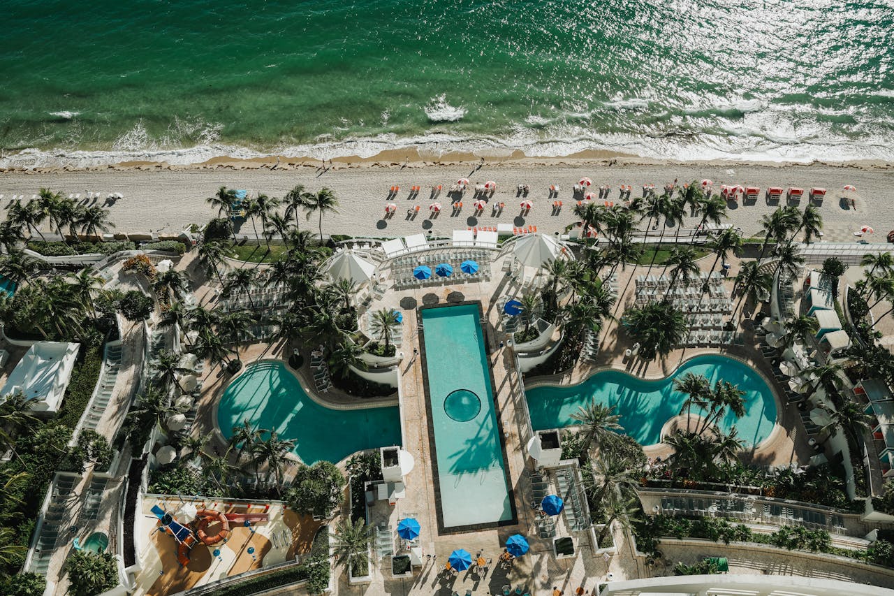 Aerial view of a beachfront resort with pools, palm trees, and sandy beach along the turquoise sea.